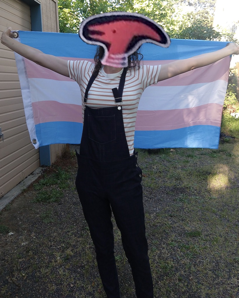 Me, wearing blue overalls, holding a trans pride flag